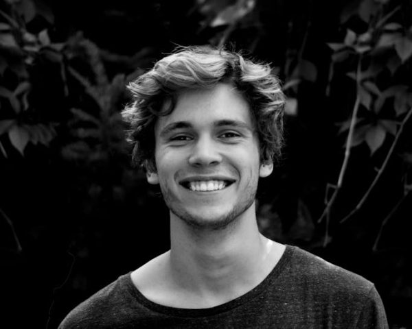 A black and white portrait of a young man smiling outdoors with natural background.